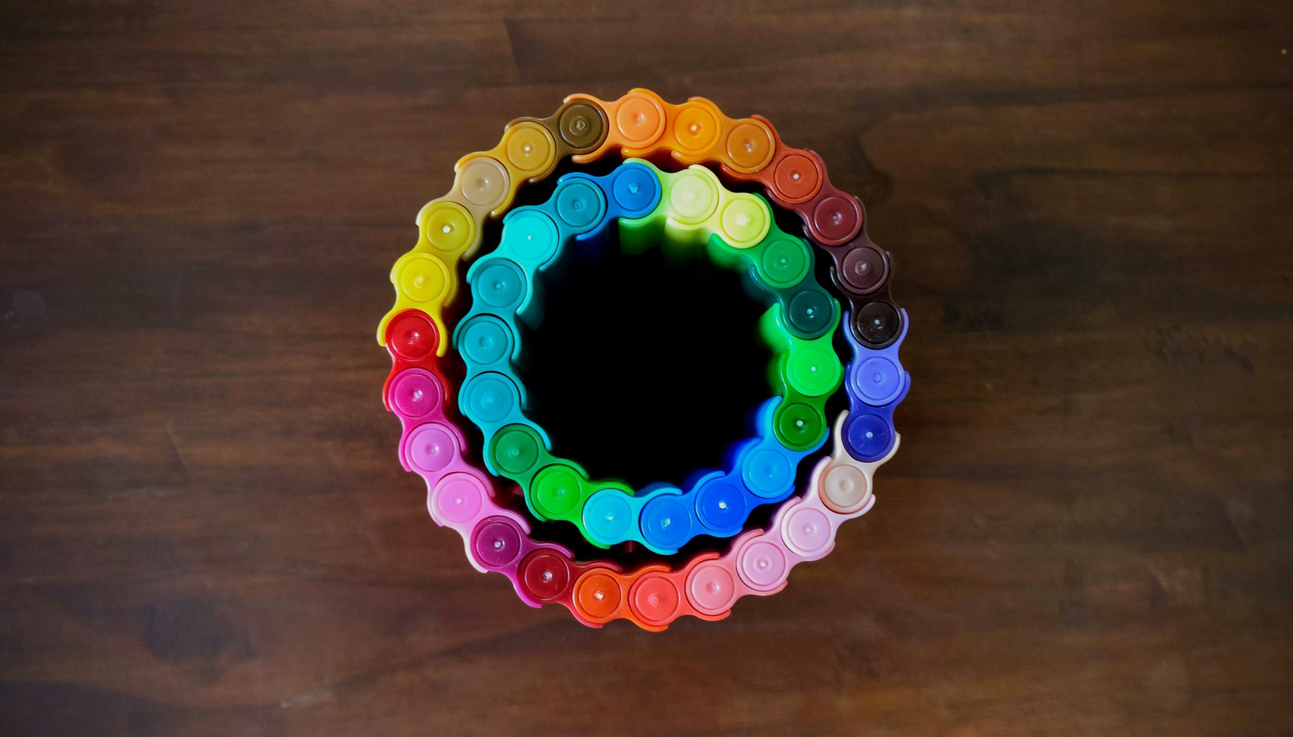Different colour markers connected together in circles. The image is photographed from above so only the tops of the lids are visible. The table they're on is wood. It looks like two rainbow circles, one inside the other.