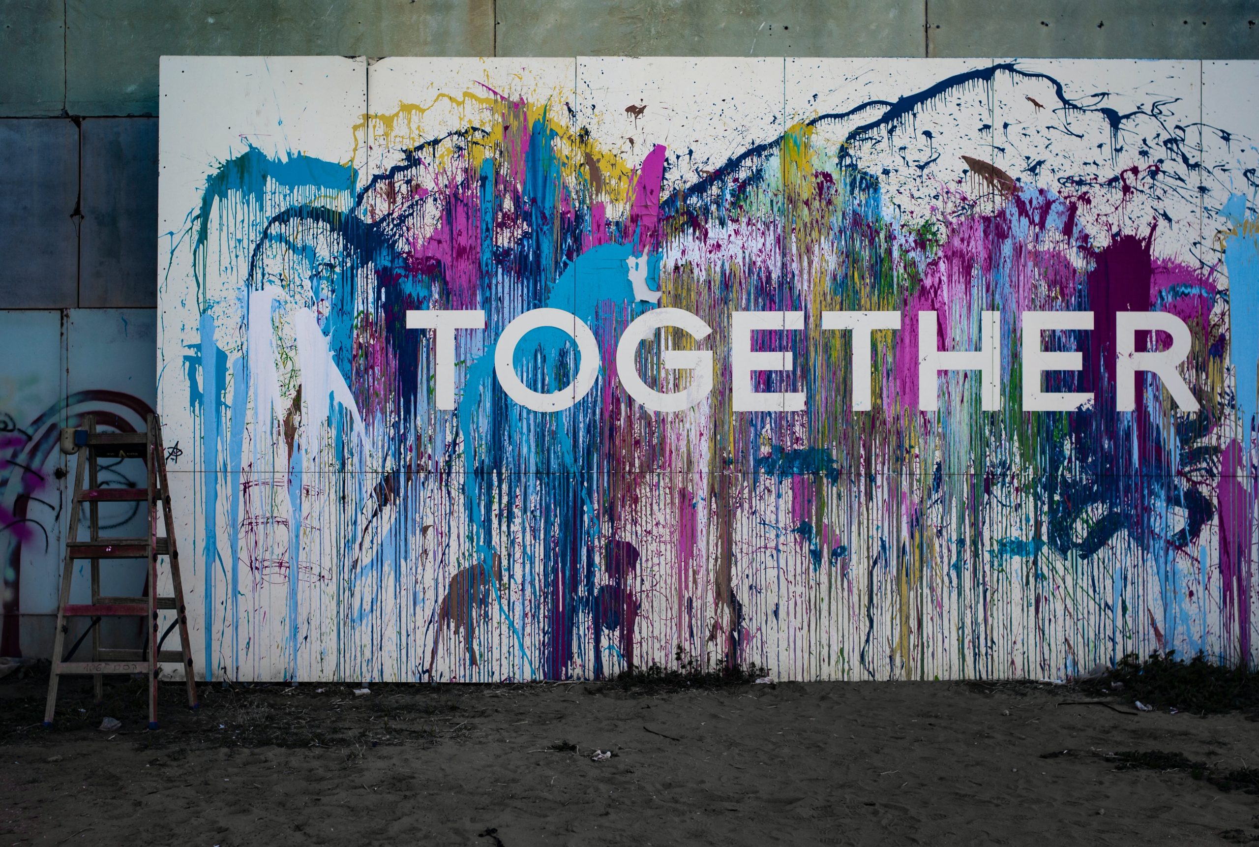 A large white canvas leaning against a wall made of grey bricks splashed with different colours of pain with the word Together across it in white letters.