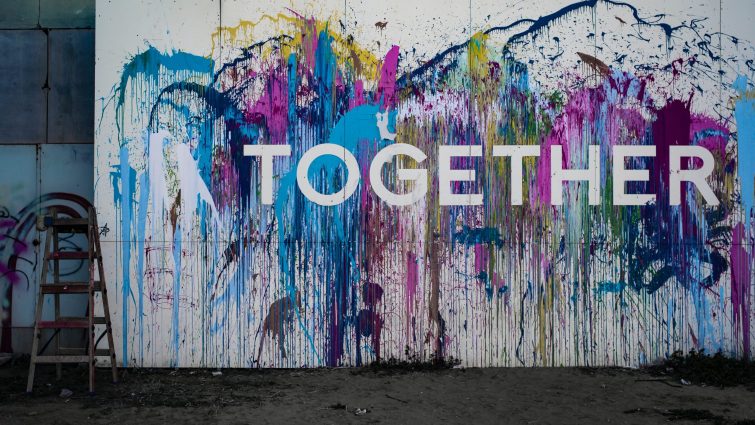 A large white canvas leaning against a wall made of grey bricks splashed with different colours of pain with the word Together across it in white letters.